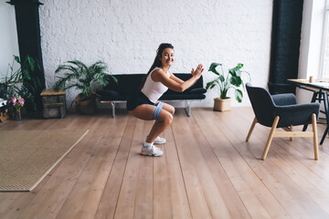 Fototapeta premium Sporty woman doing resistance band squats in bright apartment, combining wellness with personal discipline and digital-free self-care practice.