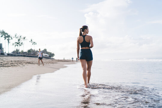 Young brunette woman walks barefoot through shallow beach water, wearing sportswear and earphones, embodying digital solitude, health focus, and unplugged self-care.