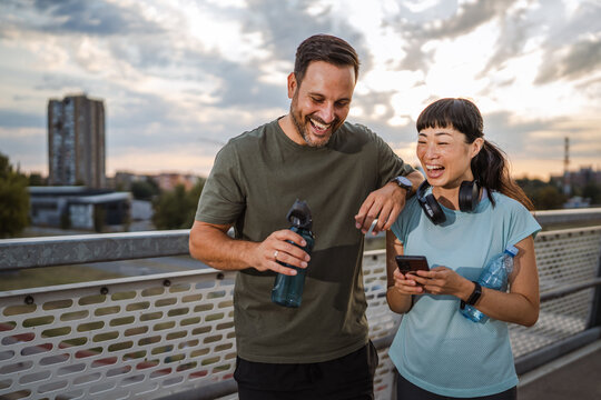 Diverse couple laughing and checking smart phone after urban exercise