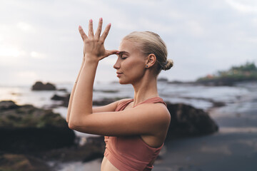 Soft close-up of woman practicing yoga mudra with fingers to third eye, eyes shut, standing against ocean at dusk, symbolizing personal connection, spiritual vision and post-digital awareness.