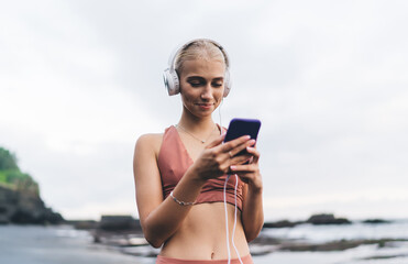 Blonde woman smiles softly at phone screen, wearing headphones, embodying joy, empowerment, and the modern relationship between body, emotion and smart digital connection.