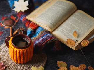 knitted orange cup of hot coffee on a table with a colorful warm scarf and an old book. Autumn composition
