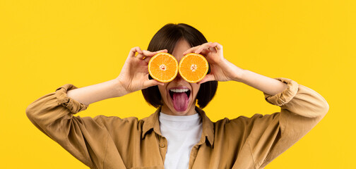 Funny young lady covering eyes with orange halves, showing tongue, fooling around over yellow studio background. Young woman placing oranges over her eyes, having fun