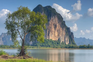 Naklejka premium Lush landscape with a lone tree by a calm river, a dramatic limestone mountain in the background