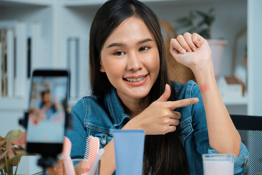 Young beautiful Asian showing cosmetic makeup lipstick matte pastel color testing in bestseller collection on arm test promotion on social media online recording smartphone at modern room. Stratagem.