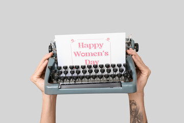 Hands holding vintage typewriter and festive postcard with text HAPPY WOMEN'S DAY on grey background