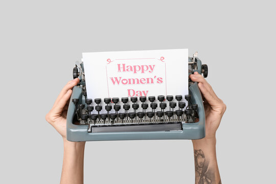Hands holding vintage typewriter and festive postcard with text HAPPY WOMEN'S DAY on grey background - Powered by Adobe