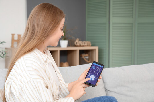 Young woman using mobile phone with smart home security system control panel on sofa in room