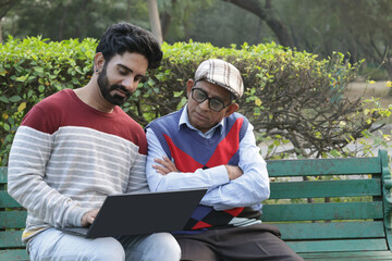 Fototapeta premium Indian Young son working on laptop for helping father related online works at park. Concept : Online ticket, travel payment, visa apply, withdraw online pension payment and online booking air tickets.