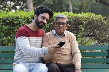 Fototapeta premium Indian Handsome son sharing smart phone with father at park. Concept : Digital India and mobile banking