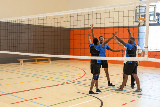 Diverse male volleyball players in blue jerseys raising arms in gym near net, copy space
