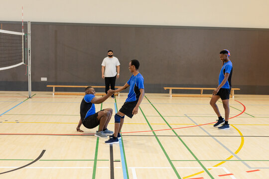 Diverse male volleyball team helping teammate up on volleyball court with knee pad and net post
