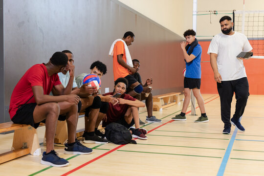 Diverse male volleyball team on wooden bench holding volleyballs in gym while coach walking with tab