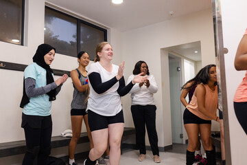 Diverse female players standing and clapping in locker room around bench with coat hooks mirror