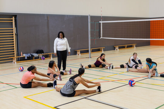 Diverse female volleyball players sitting on court floor stretching hamstrings under volleyball net
