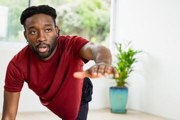 African American man holding plank with arm extending forward at home in red shirt by window