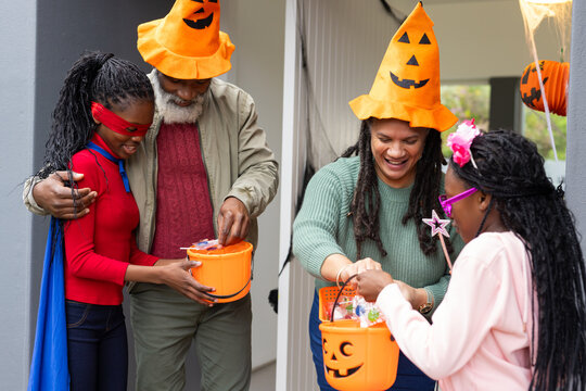Diverse hosts wearing pumpkin hats placing candy into pumpkin buckets on porch with spiderweb