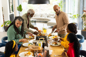 Diverse extended family sitting at wooden table in living room sharing eggs toast and orange juice