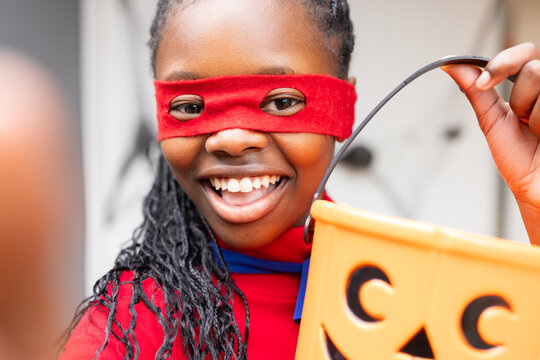 African American girl smiling and holding jack-o'-lantern candy bucket on porch wearing red mask
