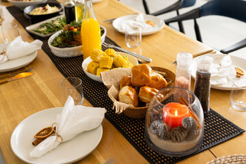 Black runner is sitting on wooden table with bread rolls, corn, orange juice bottle, lit candle