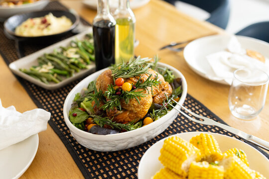 Roasted chicken is resting in casserole dish on wooden table with corn, beans and mashed potatoes