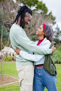 Diverse couple hugging on grassy lawn in park wearing denim jacket and holding green sweater