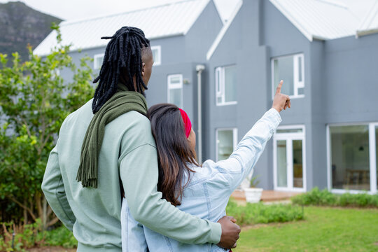 Diverse couple standing on grassy lawn before grey two-story house pointing toward mountain slope