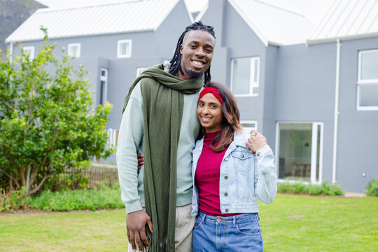Diverse couple smiling while standing on manicured lawn facing townhouse fa&ccedil;ades with picket fence