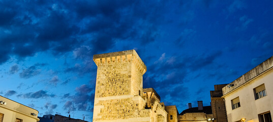Torre histórica de piedra que se alza en el centro de una plaza al anochecer.  El cielo es de un...
