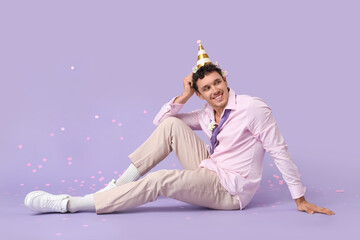 Young man in party hat with flowers sitting on lilac background