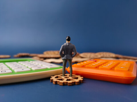 The image depicts a miniature figure of a businessman standing on a wooden gear, with calculators in the foreground