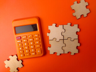 The image displays a small orange electronic calculator alongside four interlocking wooden puzzle pieces on an orange background