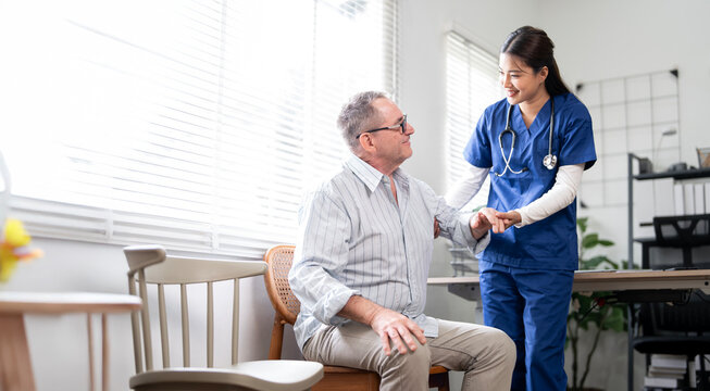 A friendly female nurse in blue scrubs stands beside an elderly male patient, offering him comfort and support. The clinic or home setting suggests a moment of compassionate healthcare,