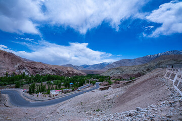 scenic view of a winding mountain road cutting through a dry rocky landscape with lush green trees nestled in the valley below, set against a dramatic blue sky with scattered clouds and distant snow