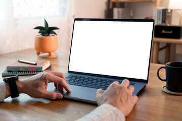 A person's hands are visible as they work on a modern laptop with a blank, white screen. The setting is a cozy, wooden desk, suggesting work-from-home