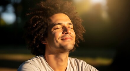 A man with curly hair, eyes closed, and a peaceful expression enjoys the golden sunlight outdoors.