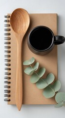 Minimalist flat lay featuring a wooden spoon, black mug, journal, & eucalyptus branch on a white background. A cozy, simple aesthetic