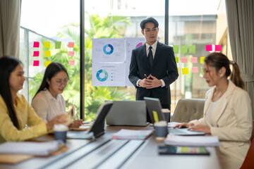 Asian businessman leading meeting with team in modern office using sticky notes and pie charts