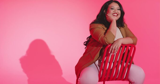 Woman straddling red metal chair in studio, resting chin, brushing hair, leaning, copy space