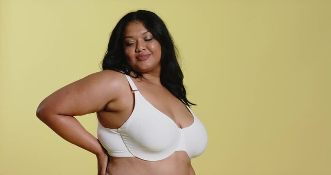 Woman adjusting white bra strap for better fit and standing, posing in studio on yellow backdrop