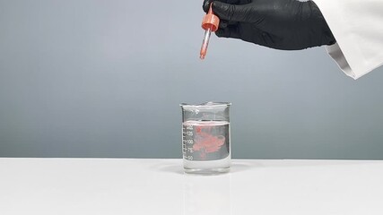 Scientist hand in black nitrile glove dropping red liquid from pipette dropper into glass beaker of water, laboratory experiment on gray background - Powered by Adobe
