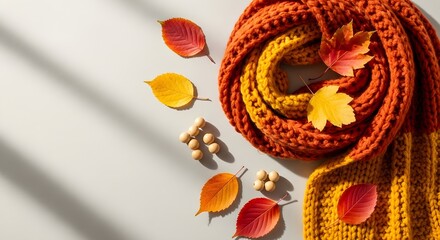 Fototapeta premium Cozy autumn scene with a warm knitted scarf, colorful fallen leaves, and natural light casting shadows on a neutral background.