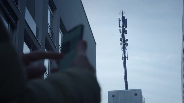 An individual is checking their smartphone while a cell tower looms in the background
