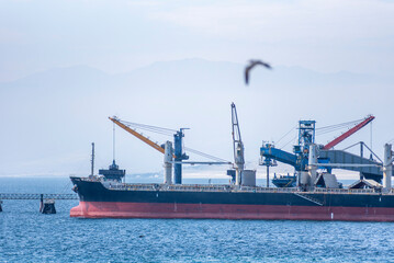 Fototapeta premium Cargo ship moored at Mejillones port, Chile, alongside a long pier. The vessel is being loaded via a conveyor belt system connected to the pier.