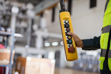 Close-up of worker operating industrial crane control pendant with emergency stop button in factory. Focus on safety, heavy equipment handling, and industrial operations