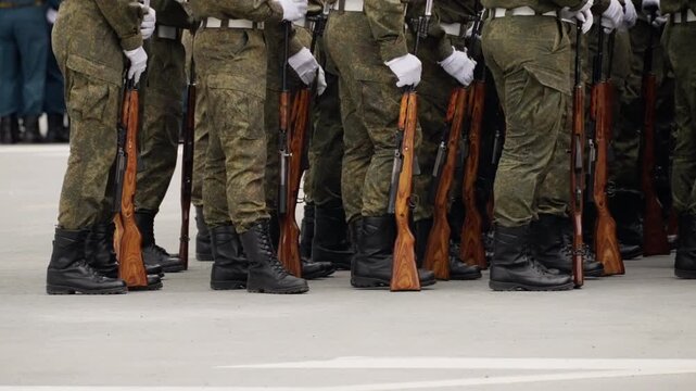 Close view of uniformed soldiers in tight formation with wooden-stock rifles and bayonets grounded, wearing camouflage fatigues, black boots, and white gloves during a drill