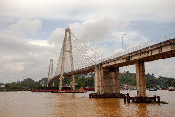 The magnificent Mahkota II Bridge spanning the Mahakam River in Samarinda, Borneo. A tugboat pulls a coal barge underneath, showcasing the river's vital role for industrial transport.