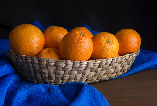 Still life photography of oranges in a hand-woven basket. A blue tablecloth on a wooden table with a dark background. horizontal