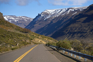 Asphalt road extending through an Andean Patagonian landscape with grass, bushes, rocky hills, and snow-covered mountains, symbolizing adventure and journey during an amazing roadtrip.
