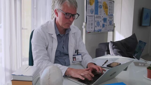 Focused senior doctor in glasses and white coat typing on laptop in modern medical office. Healthcare professional working with documents and digital records at desk.